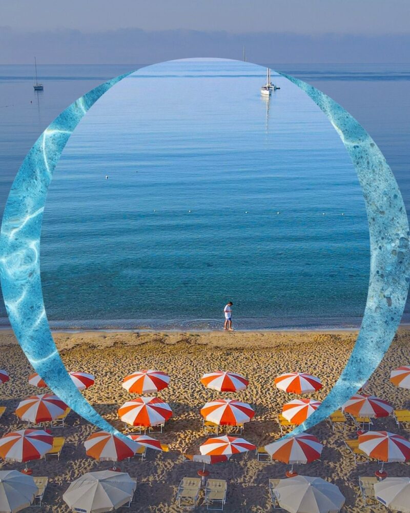 Spiaggia con ombrelloni rossi e bianchi, mare calmo, barche e una persona che cammina sulla riva.